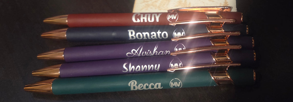 Five custom-engraved ballpoint pens in different colors (red, navy, purple, and green) with rose gold metal accents, stacked horizontally on a dark surface.