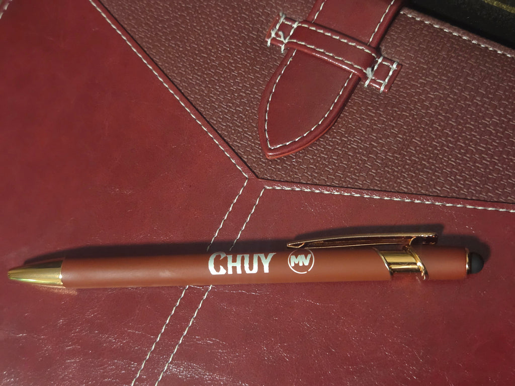 A brown ballpoint pen with gold accents and white CHUY branding, resting diagonally on a textured reddish-brown leather surface with white stitching.