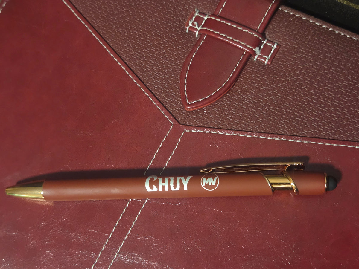 A brown ballpoint pen with gold accents and white CHUY branding, resting diagonally on a textured reddish-brown leather surface with white stitching.