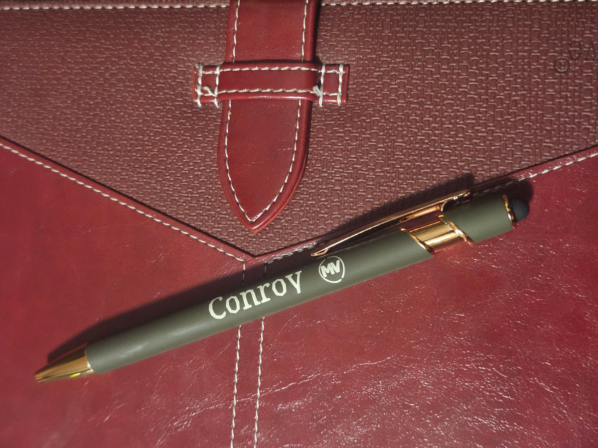 A dark olive green Conroy ballpoint pen with gold accents and white logo printing, resting diagonally on a textured dark red leather surface next to a matching red leather strap.